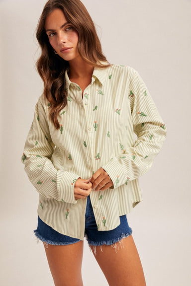 Woman wearing a striped button up shirt with cactus embroidery and denim shorts on a plain background