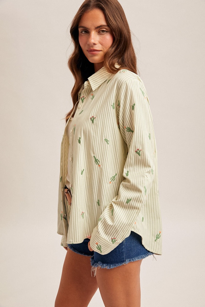 Woman wearing a striped button up shirt with cactus embroidery and denim shorts on a plain background