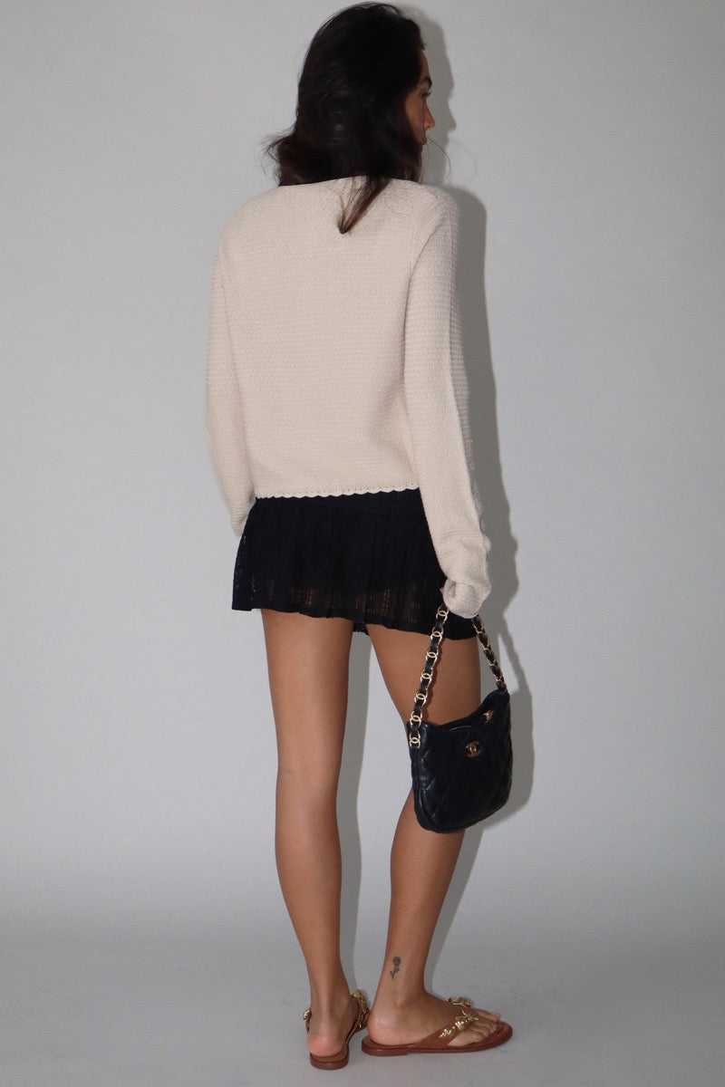 Back view of a woman wearing a beige cardigan with gold shell buttons and a black skirt against a white background