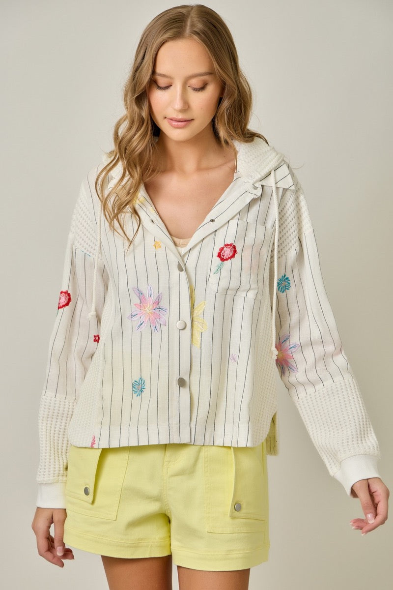 Woman wearing a light-colored embroidered jacket with colorful designs and yellow shorts on a neutral background