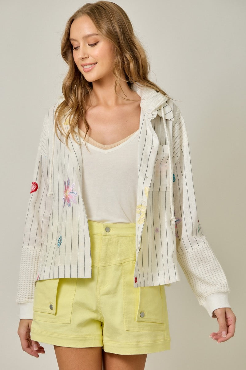 Woman wearing a light-colored jacket with embroidered designs and yellow shorts on a neutral background